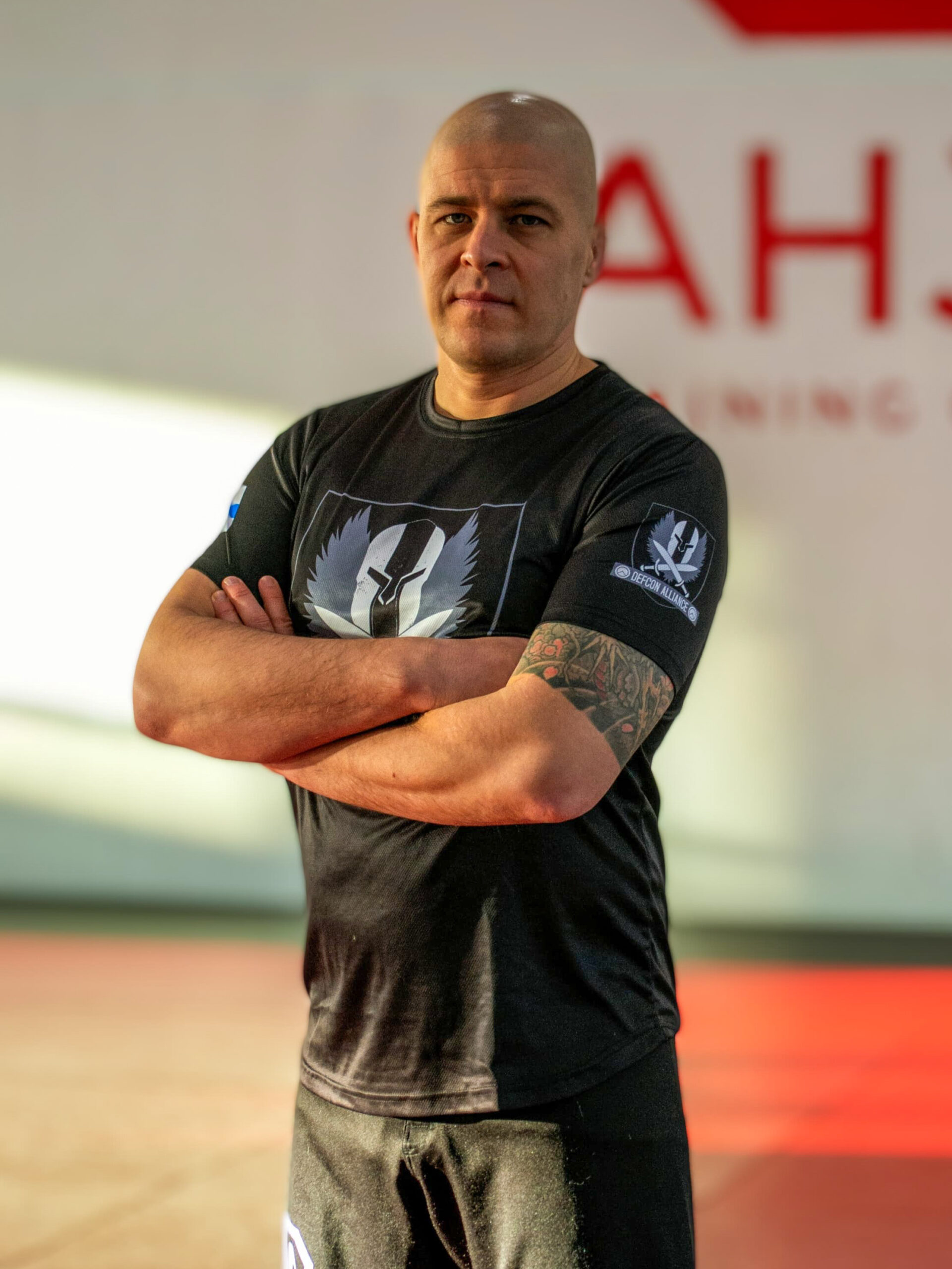 Valmentaja Tomi Jaakkola seisoo käsivarret ristissä Defcon alliancen paita päällä ja katsoo kameraan. Taustalla on Ahjo Training Centerin puna-musta tatami ja Ahjon logo.